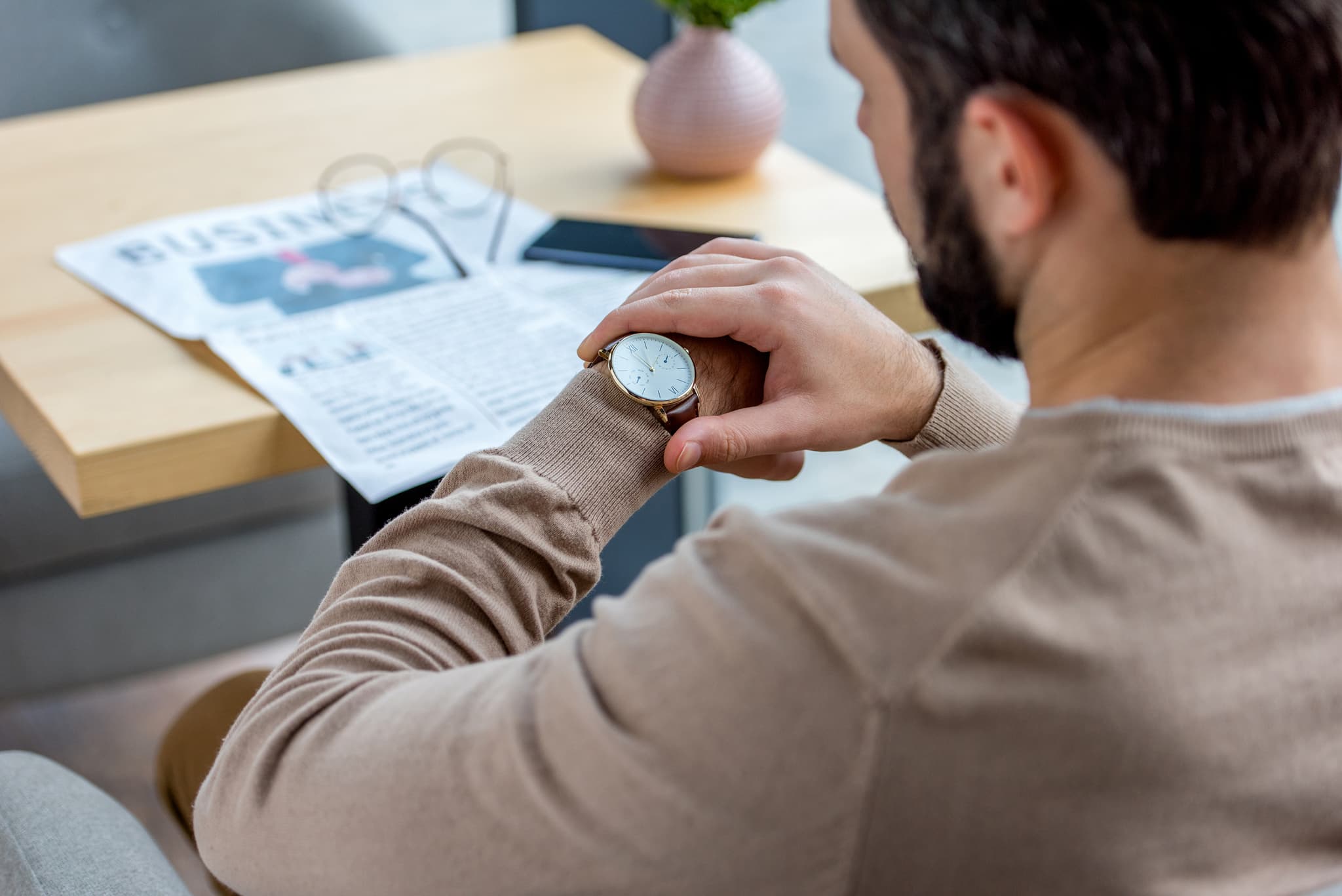 A bearded man at a desk checking his wristwatch over a business newspaper