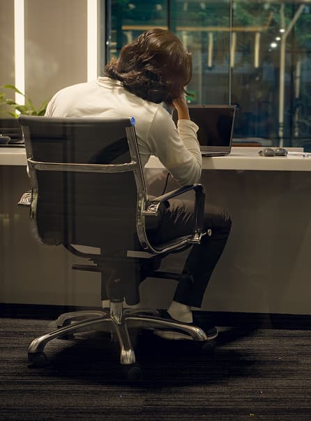 A person slumped in an office chair at night, back turned to a laptop
